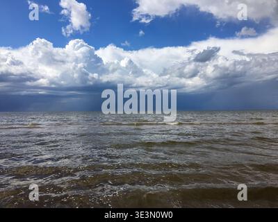 Wellen Rollen sanft zum Ufer unter einem hellblauen Himmel voller weißer Wolken. Die Sonne wirft Licht auf die Wasseroberfläche. Die Szene zeigt das Weite Stockfoto
