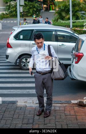 Ein Mann geht über die Straße und hält Essen und Tasche. Städtische Umgebung mit geparkten Autos und Quersteg. Freizeitkleidung, konzentriert auf sein Essen. Im Hintergrund sind Pads zu sehen Stockfoto