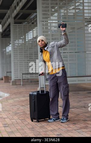 Mann, der am Abholbereich steht und den Koffer hält, das Smartphone hochhebt, Beanie und Kopfhörer trägt. Reisende, Pendler, Gepäck, Telefon, Stadt, Transit, s Stockfoto