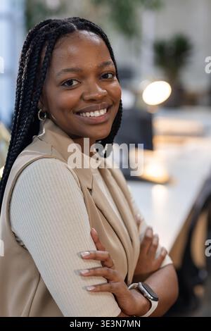 Afroamerikanerin lächelt mit überkreuzten Armen im Büro und trägt Blazer, Smartwatch bei Schreibtischlampe Stockfoto