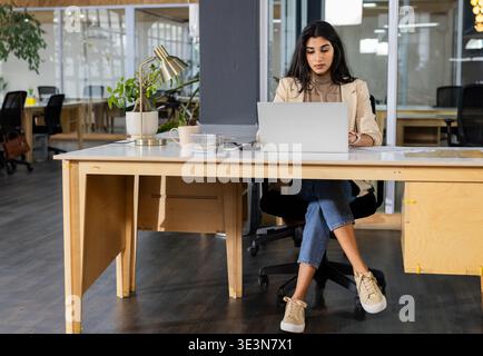 Indische Frau, die an einem Holztisch in einem offenen Büro mit silbernem Laptop und einer Messingtischlampe arbeitet Stockfoto
