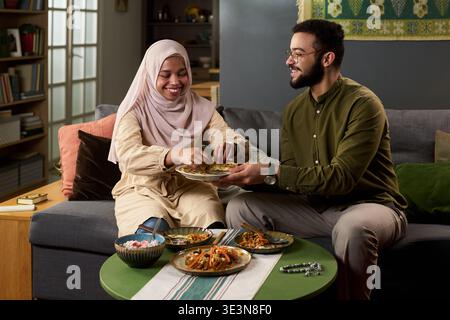 Junge Erwachsene muslimische Frau und junger erwachsener muslimischer Mann sitzen auf dem Sofa und teilen gemeinsam Mahlzeiten, lächelnd und interagierend, traditionelle Gerichte auf dem Tisch Stockfoto