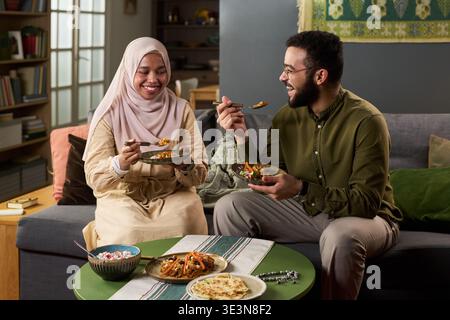 Junge Erwachsene muslimische Frau im Hijab und ihr Mann sitzt auf dem Sofa und isst gemeinsam traditionelle Mahlzeiten, lächelnd und interagierend, während sie Schüsseln hält Stockfoto