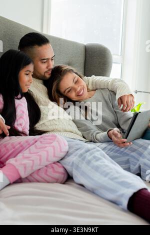 Vielfältige Familienkuscheln auf Polsterbett im hellen Schlafzimmer mit Tablet mit gemusterter Decke Stockfoto