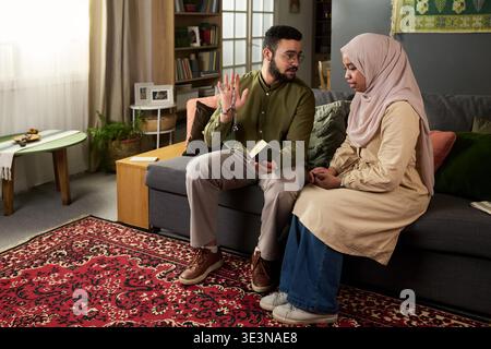 Muslimischer Mann mittleren Alters, der Bücher hält, während er auf dem Sofa neben einer jungen erwachsenen muslimischen Frau sitzt und aufmerksam im Wohnzimmer zuhört Stockfoto