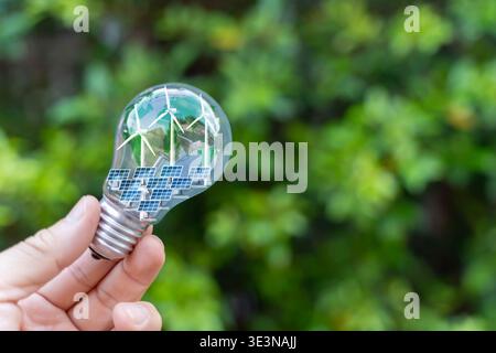 Nahaufnahme einer Hand, die eine Glühbirne mit Windturbinen, Solarpaneelen und einer Kugel im Inneren auf grünem Naturhintergrund hält, die erneuerbare Energie symbolisiert, Stockfoto