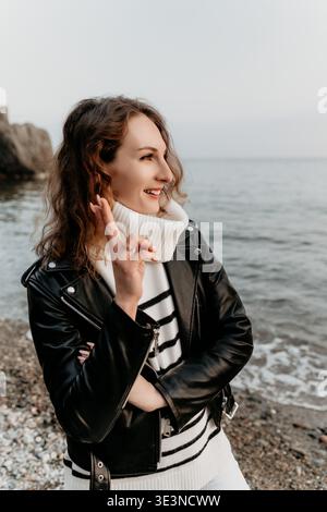 Frau Lederjacke Strand lächelnde Frau mit gekreuzten Fingern steht am Kieselstrand in gestreiftem Pullover und schwarzem Mantel Stockfoto