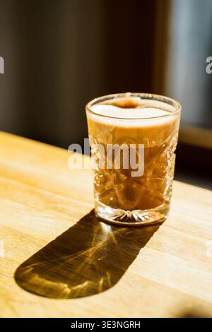 Heißer Kakao im Glas mit cremigem Schaum, Sonnenlicht wirft Schatten auf Holztisch, gemütliche Café-Atmosphäre, warmes Getränk, Komfortkonzept Stockfoto