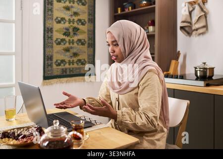 Junge Erwachsene muslimische Frau im Hijab, die am Küchentisch sitzt und Gesten beim Videoanruf auf dem Laptop mit Tee und Snacks auf dem Tisch zu Hause macht Stockfoto