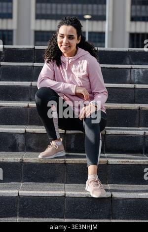 Frau, die auf Steintreppen vor dem modernen Gebäude sitzt, lächelt und eine Jacke mit Smartwatch trägt Stockfoto