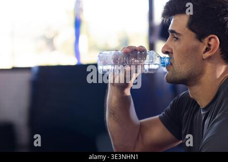 Mann trinkt aus einer klaren Plastikflasche, trägt dunkelgraues athletisches T-Shirt im Fitnessstudio, Kopierraum. Hydratation, Hintergrundbeleuchtung, Behälter, Kondensation, vorn Stockfoto