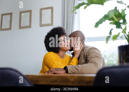 Verschiedene Paare sitzen zu Hause Esstisch mit gelbem Pullover und teilen sich zärtlichen Kuss neben der Pflanze Stockfoto