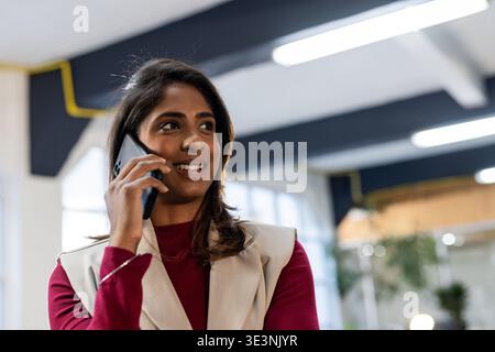 Indische asiatische Frau mit kastanienbraunem Oberteil und beigefarbener Weste, die im Großraumbüro mit Smartphone spricht Stockfoto