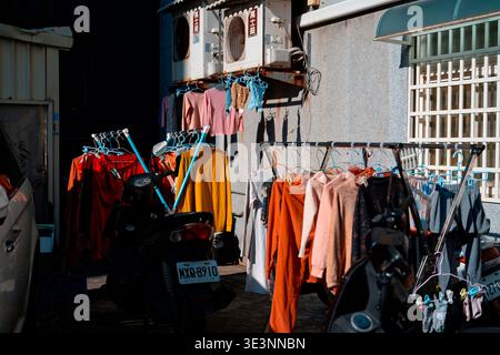 Taoyuan, Taiwan - 22. Januar 2023: Farbenfrohe Kleidung hing an Regalen im Freien neben Rollern bei hellem Sonnenlicht vor einem traditionellen taiwanesischen Bewohner Stockfoto
