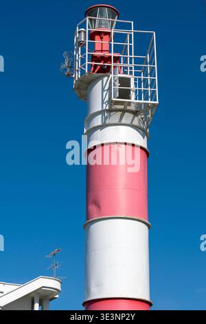 Ein rot-weißer Leuchtturm mit Metallgeländer und Leuchtfeuer, der hoch vor einem leuchtend blauen Himmel steht und die Seeschifffahrt, die Küstensicherheit, Stockfoto