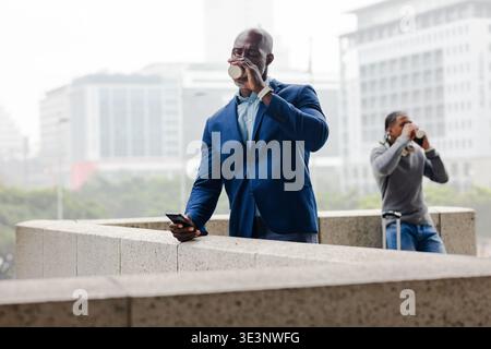 afroamerikanischer Mann mit Anzug, der auf der Terrasse steht und Kaffee mit Smartphone trinkt Stockfoto