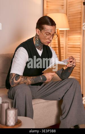 Ein hübscher Mann mit Tattoos und stilvoller Brille konzentriert sich auf ein Buch, während er sich auf einem Sofa entspannt. Stockfoto