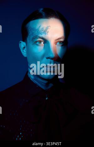 Der junge Mann zeichnet sich durch auffällige Merkmale und Stil aus, die durch intensives blaues Licht verstärkt werden. Stockfoto