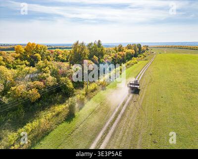 Sonnige Luftaufnahme eines landwirtschaftlichen Traktors, der Anhänger auf staubigen Wegen in der Nähe von grünem Feld und gelbem Wald zieht und ländliche Arbeit zeigt, selektiver Fokus Stockfoto