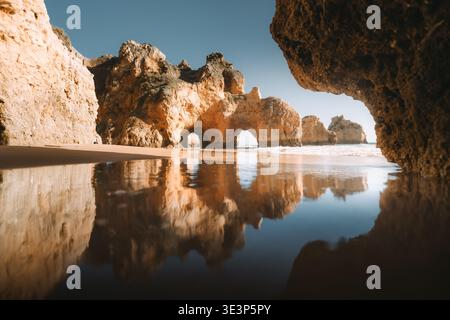 Blick auf goldene Klippen, die sich im ruhigen Wasser spiegeln, während Wellen sanft das Ufer in der Algarve, Alvor, Faro, Portugal, küssen. Stockfoto