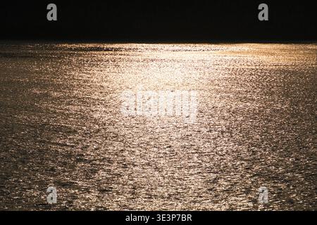 Ruhige Küstenlandschaft bei Sonnenuntergang mit hellem goldenem Sonnenlicht, das sich über die ruhige Wasseroberfläche reflektiert, dunkle Silhouette eines bewaldeten Hügels auf dem Ho Stockfoto