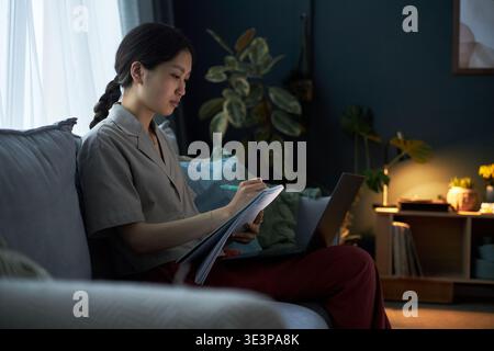 Asiatische junge Erwachsene Frau, die auf dem Sofa sitzt und im Notebook schreibt, während sie einen Laptop benutzt, konzentriert sich auf das Lernen oder Arbeiten in einem modernen Wohnzimmer mit Pflanzen und weichem Tageslicht Stockfoto
