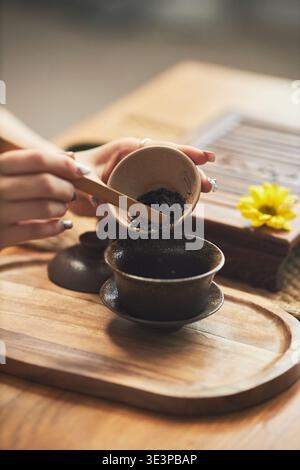 Asiatische junge Erwachsene Frau, die traditionelle Teezeremonie vorbereitet, eine Keramikschale hält und mit einem Holzlöffel lose Teeblätter in die Teekanne gießt, ein Holztablett und eine sichtbare Blume Stockfoto
