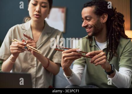 Asiatische junge Erwachsene Frau demonstriert Essstäbchen Technik zu lächelndem jungen schwarzen Erwachsenen Mann, beide sitzen drinnen und nehmen gemeinsam kulturelle Lernaktivitäten wahr Stockfoto