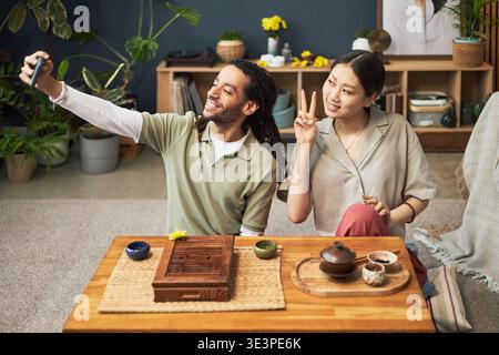 Junge Erwachsene asiatische Frau und junger Erwachsener Schwarzer Mann sitzen zusammen am niedrigen Tisch und machen Selfie mit Smartphone, asiatische Frau macht Friedenszeichen Geste, traditioneller Tee auf Tisch Stockfoto