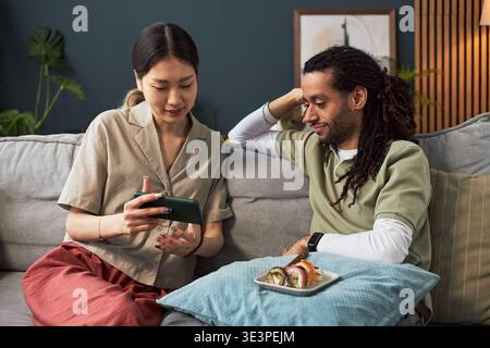 Asiatische junge Erwachsene Frau zeigt Smartphone zu Schwarzen jungen Erwachsenen Mann, der zusammen auf Sofa sitzt, sowohl lächelnd als auch interagierend, Sushi-Teller auf Kopfkissen vor der Tür, zwangloses Zuhause Stockfoto