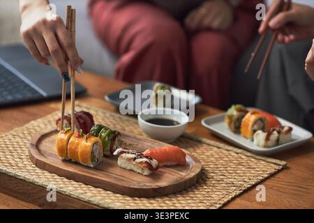 Asiatische junge Erwachsene pflücken verschiedene Sushi-Brötchen aus Holzplatte, teilen traditionelle japanische Gerichte während einer ungezwungenen Mahlzeit, Sojasauce in einem kleinen Gericht auf dem Tisch Stockfoto