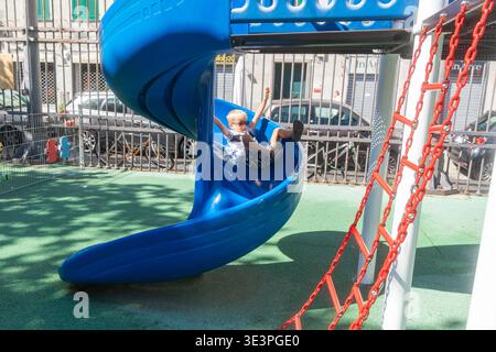 Kind rutscht an sonnigen Tagen auf dem blauen Spielplatz im Stadtpark hinunter. Aktive Kindheitsmomente mit Outdoor-Spaß, Bewegung und gesundem Spiel. Stockfoto