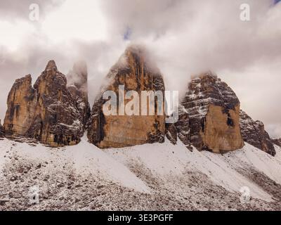 Aus der Vogelperspektive auf die zerklüfteten Gipfel des Tre Cime di Lavaredo, die durch schneebedeckte Wolken durchdringen, Tre Cime di Lavaredo, Venetien, Italien. Stockfoto