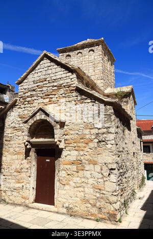 Das Äußere der historischen Steinkirche, die in hellem Sonnenlicht getaucht ist, zeigt mittelalterliche Architektur, strukturierte Kalksteinwände und eine rustikale Holztür unter einem leuchtend blauen Himmel. Ruhige, zeitlose Atmosphäre in einem schmalen Innenhof. Stockfoto