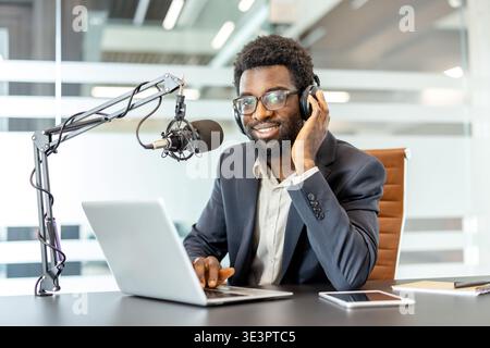 Junger Mann mit Kopfhörern und Brillen, der selbstbewusst in ein Mikrofon spricht, während er einen Laptop benutzt, und ein Audioprogramm eines Berufs sendet Stockfoto