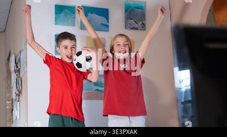 Aufgeregte Jungs in roten Hemden, die mit erhobenen Armen anfeuern, während sie zu Hause ein Fußballspiel im fernsehen anschauen Stockfoto