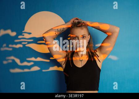 Frau mit Dreadlocks und Tattoos, die mit Händen auf dem Kopf posiert, mit kreativer Lichtprojektion Stockfoto