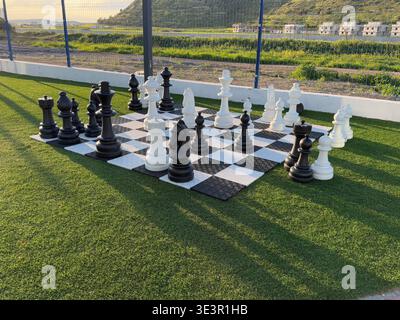 Riesiges Schach im Freien auf einem grasbewachsenen Feld mit malerischen Bergen und sonnendurchflutetem Himmel im Hintergrund. Mobiles Foto Stockfoto