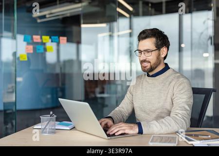 Junger, professioneller Mann mit Brille lächelt, während er sich an einem modernen Schreibtisch auf sein Notebook konzentriert, und vermittelt Produktivität, Selbstvertrauen und eine komfortable, intelligent-lässige Arbeitsumgebung Stockfoto