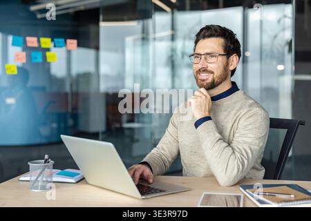 Glücklicher, bärtiger professioneller Mann mit Brille, lächelnd am Schreibtisch, mit einem Laptop und wegschauend, über neue Geschäftsideen in einem kreativen Büroarbeitsplatz nachdenken Stockfoto