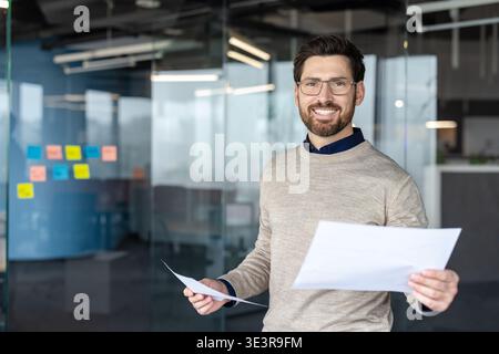 Junger Geschäftsmann mit Brille und Pullover, der selbstbewusst lächelt, während er Papiere mit einem hellen, professionellen Büro-Innenraum als Hintergrund hält Stockfoto