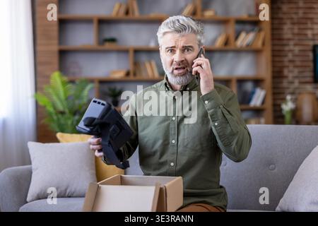 Reifer Mann, der ein beschädigtes Virtual-Reality-Headset von einer Online-Lieferung erhält und frustriert aussieht, als er mit dem Kundenservice in seinem Wohnzimmer über ein Mobiltelefon sprach Stockfoto