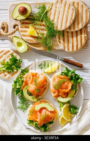 Gegrillte Pita-Sandwiches mit dünn geschnittenem Räucherlachs, Avocado, Dill und Zitronenscheiben, serviert auf einem weißen Teller auf weißem Holztisch mit Messer, flach Stockfoto