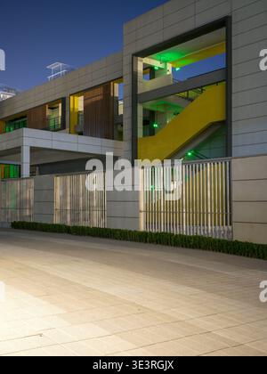 Ein leerer gepflasterter außenplatz führt zu einem modernen Gebäude mit weißen Paneelen, gelben architektonischen Akzenten und einem Sicherheitszaun aus Metall, der bei n beleuchtet ist Stockfoto
