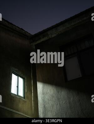 Ein flacher Blick auf eine alte verwitterte Gebäudewand mit einem einzigen hell beleuchteten Fenster, das Licht in die dunkle Nacht wirft. Die Szenenrepresse Stockfoto
