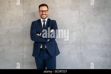 Junger glücklicher, erfolgreicher Geschäftsmann mit überkreuzten Armen lächelnd im Firmenbüro Stockfoto