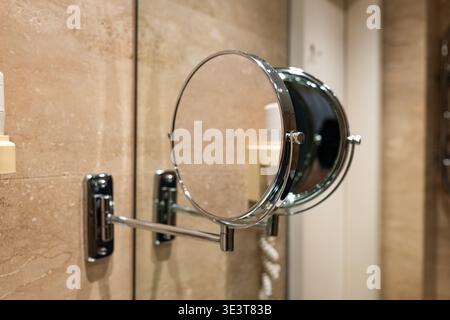 Nahaufnahme eines schlanken Schminkspiegels mit schwenkbarem Arm und poliertem silbernem Rahmen, der an einer strukturierten Steinwand in einem eleganten, modernen Badezimmer befestigt ist Stockfoto