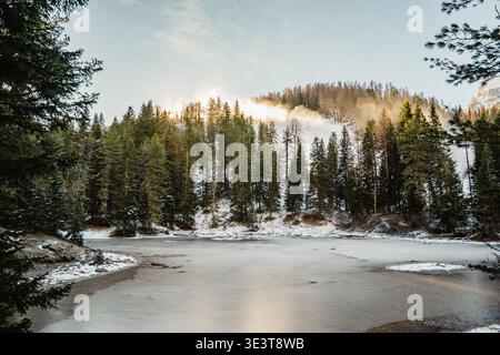 Eindrucksvoller Blick auf einen gefrorenen Bergsee mit Schnee an seinen Ufern, eingerahmt von dichten Nadelwäldern. Die niedrige Sonne filtert durch die Äste, cre Stockfoto