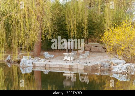 Der ruhige Teich reflektiert umliegende Bäume und Gartenelemente. Weidenbäume mit herabhängenden Ästen säumen den Wasserrand. Stühle aus Stein blicken auf den Teich in der Nähe von A Stockfoto