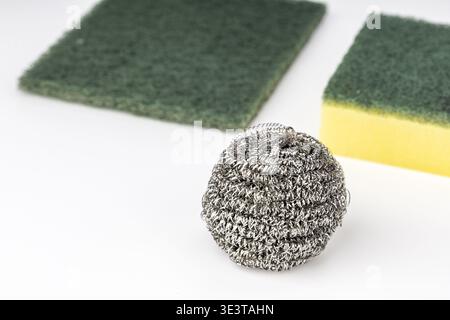 Eine glänzende Scheuerkugel aus Edelstahldrahtwolle ist im Vordergrund scharf fokussiert, mit zwei grünen Scheuerschwämmen und einer gelben Schaumküche Stockfoto